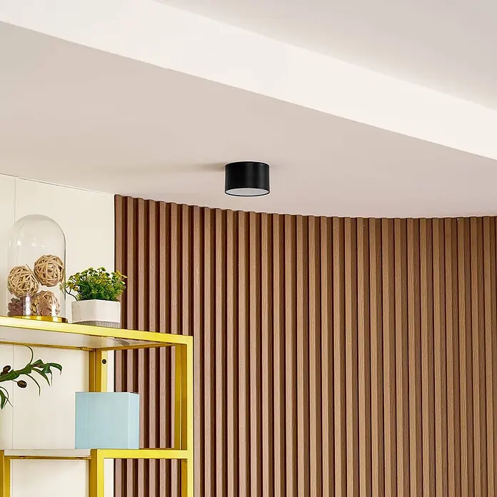 Lindby Deckenstrahler Nivoria - schwarz - Schwarze zylindrische Deckenleuchte an einer hellen Decke über einer Holzlamellenwand.