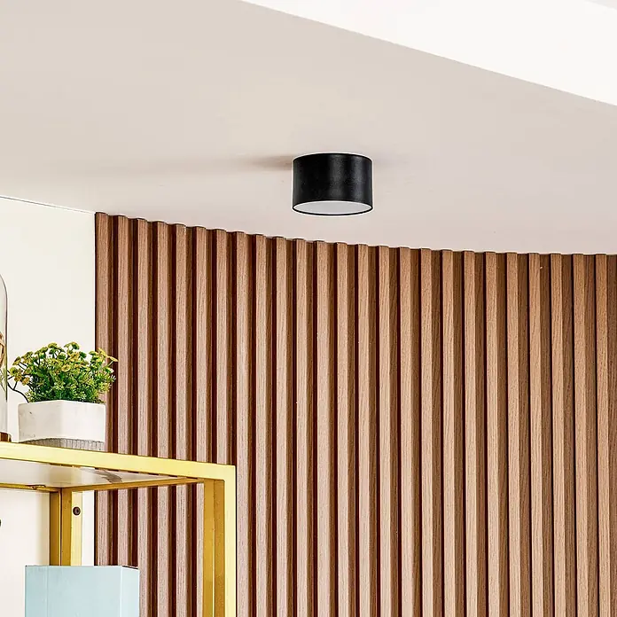 Lindby Deckenstrahler Nivoria - schwarz - Schwarze zylindrische Deckenleuchte an weißer Decke über Holzlamellenwand und goldenem Regal mit Pflanze.