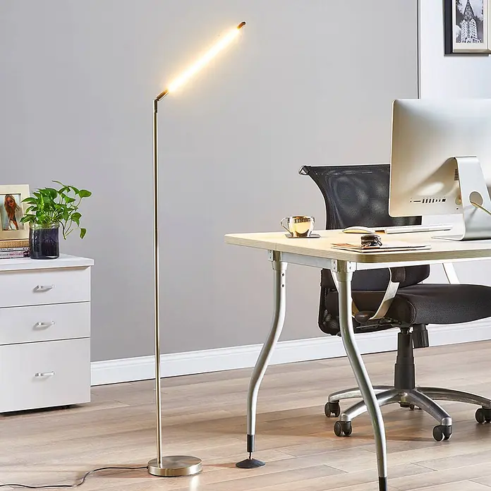 Lindby Stehleuchte Jabbo - alu / grau / zink, alu / grau / zink - Eine goldfarbene lineare LED-Stehleuchte steht neben einem hellen Schreibtisch und Bürostuhl in einem modernen Büro und spendet helles Licht.