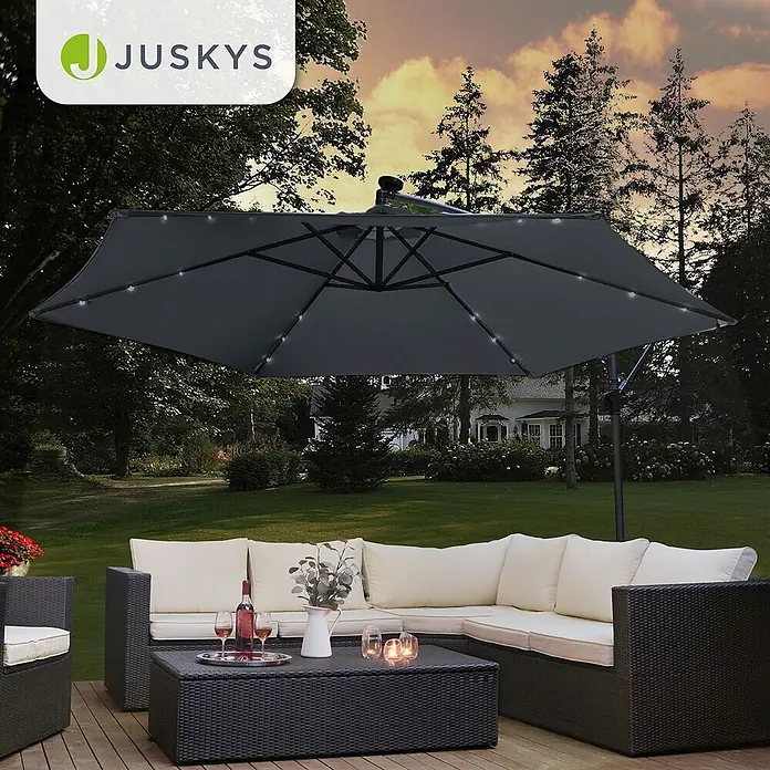 Juskys Ampelschirm rund Brazil inkl. LED - dunkelgrau, Durchmesser Bespannung: 350 cm - Schwarze Rattan-Gartenmöbel mit cremefarbenen Polstern und großem dunkelgrauen Ampelschirm mit LED-Beleuchtung auf einer Holzterrasse.