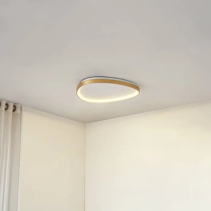 Lucande Deckenleuchte Asken - messing / gold - Ovale LED-Deckenleuchte mit goldenem Rahmen, beleuchtet eine Raumecke mit warmem Licht.
