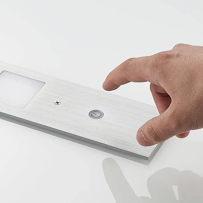 Arcchio LED-Unterbauleuchte Nortra -  - Gebürstetes silbernes LED-Lichtband mit rechteckigem Panel und Touch-Knopf, ein Finger ist dabei, ihn zu drücken.
