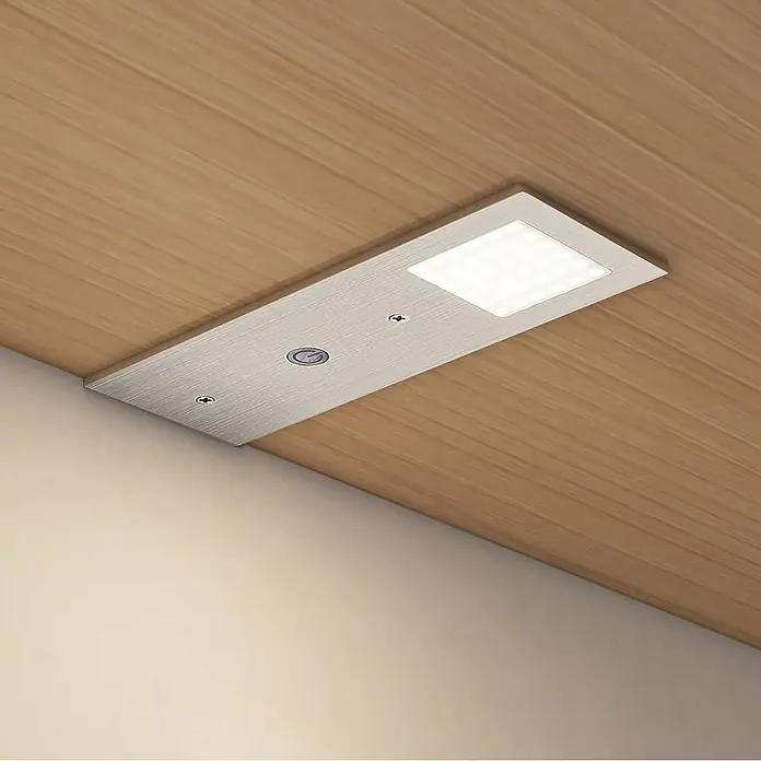 Arcchio LED-Unterbauleuchte Nortra -  - Gebürstete Metall-LED-Unterbauleuchte mit Einschaltknopf, die von einer Holzoberfläche leuchtet.