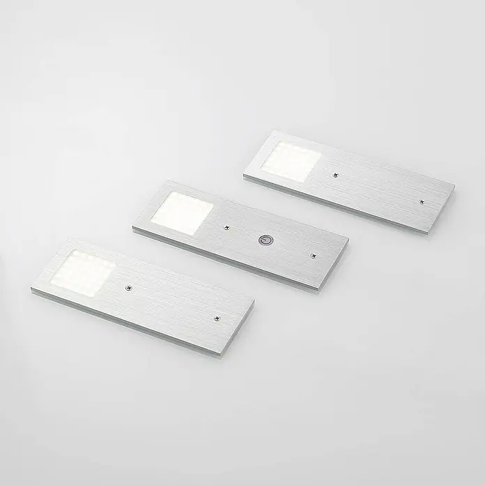 Arcchio LED-Unterbauleuchte Nortra -  - Drei rechteckige LED-Leuchten aus gebürstetem Silber mit quadratischen Lichtpaneelen. Eine Leuchte hat einen runden Einschaltknopf.