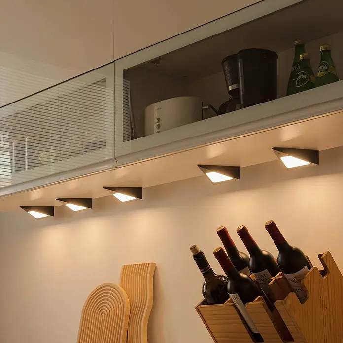 Prios LED-Unterbauleuchte Odia -  - Dunkle, dreieckige Unterbauleuchten beleuchten eine Küchenarbeitsplatte und ein Holzweinregal.