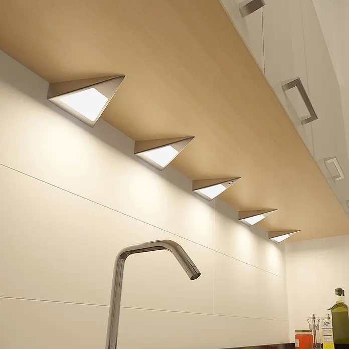 Prios LED-Unterbauleuchte Odia - L x B x H: 13,5 x 12,75 x 4 cm - Dreieckige silberne Unterbauleuchten an heller Holzfläche, die einen weißen Küchenfliesenspiegel erhellen.