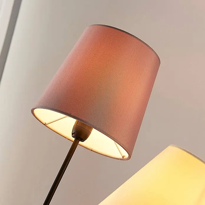 Lindby Stehleuchte Melis - creme / amber, E14 - Nahaufnahme zweier leuchtender Lampen; eine mit rötlich-braunem Stoffschirm, die andere mit cremefarbenem Stoffschirm, beide leuchtend.