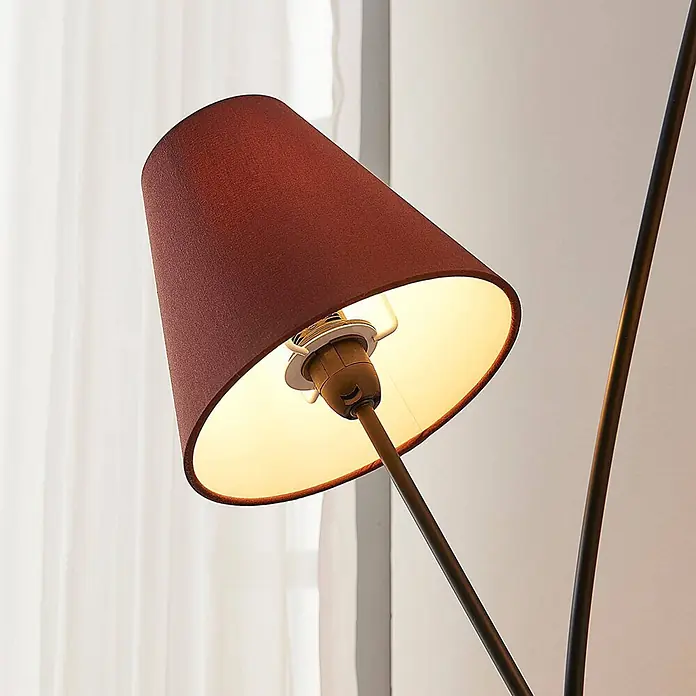Lindby Stehleuchte Melis - creme / amber, E14 - Rotbrauner Stofflampenschirm mit warmem Licht, auf dunklem Metallarm montiert, beleuchtet einen Innenraum.