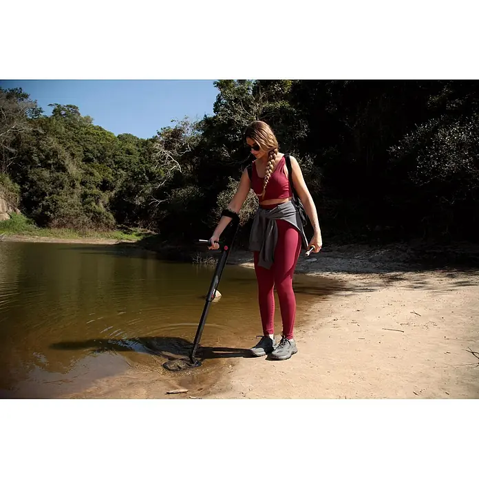 Minelab Metalldetektor GO-FIND 66 U-Light - L x B x H: 160 x 230 x 1.300 mm - Frau in roter Sportkleidung nutzt einen schwarz-roten Metalldetektor im flachen Wasser an einem Sandufer.