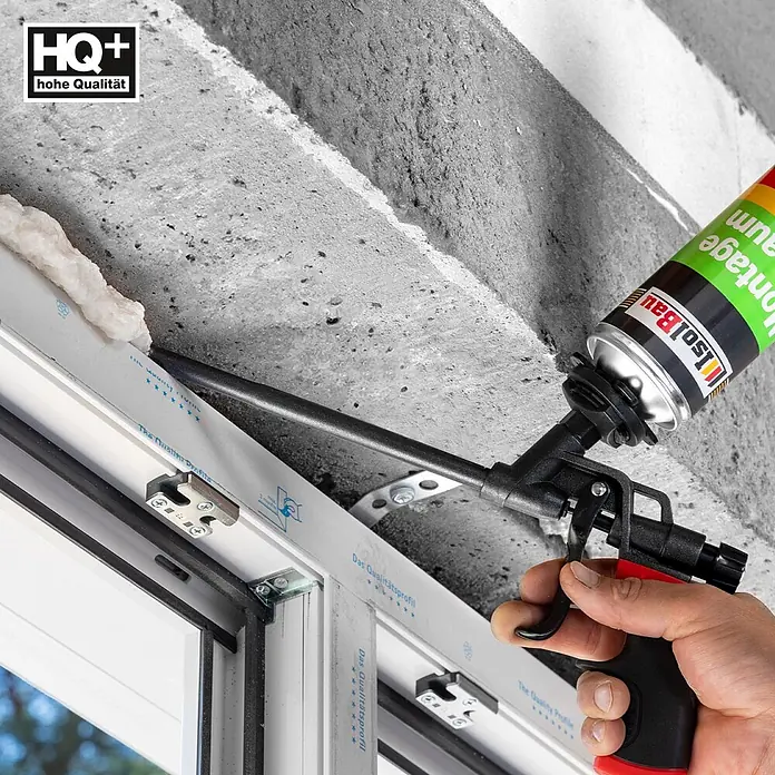 IsolBau Bauschaum 1 x Montageschaum 750 ml - 750 ml - Hand füllt mit schwarzer und roter Schaumpistole und grüner IsolBau Montageschaumdose eine Spalte am weißen Fensterrahmen vor Betonwand.
