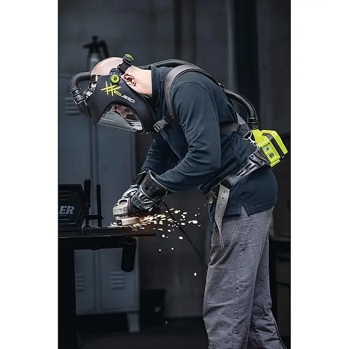 Optrel Schutzhelm Clearmaxx mit Gebläseatemschutz E3000X -  - Mann winkelschleift, trägt einen schwarzen Schweißhelm mit Visier und eine gelb-schwarze Gebläseeinheit (PAPR) auf dem Rücken.