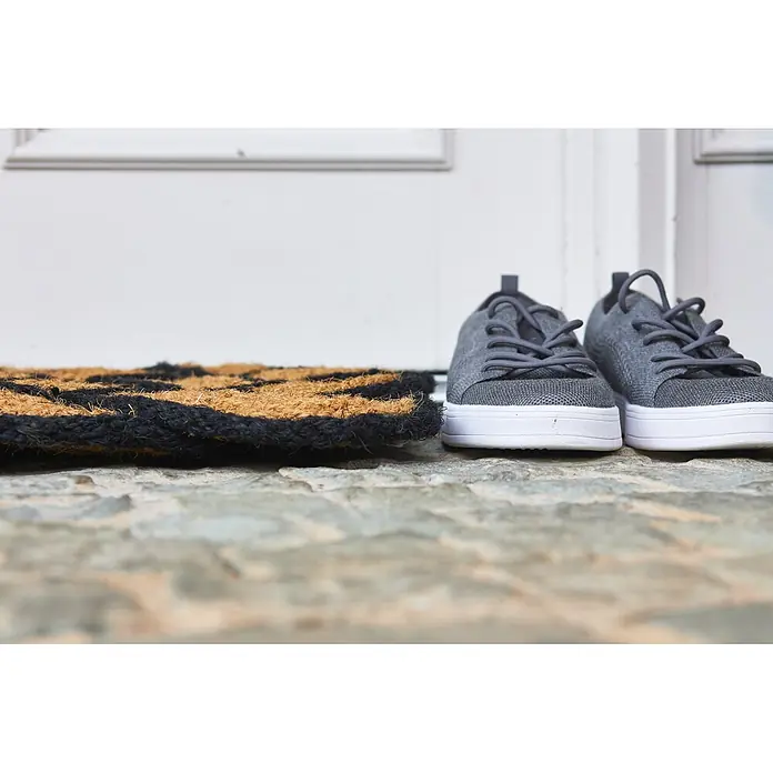 Fußmatte Nature Classic - 75 x 45 cm, Natur/Schwarz, Material Nutzschicht: 100 % Kokos - Graue Strick-Sneaker und eine gemusterte Kokos-Fußmatte auf Steinboden vor einer weißen Tür.