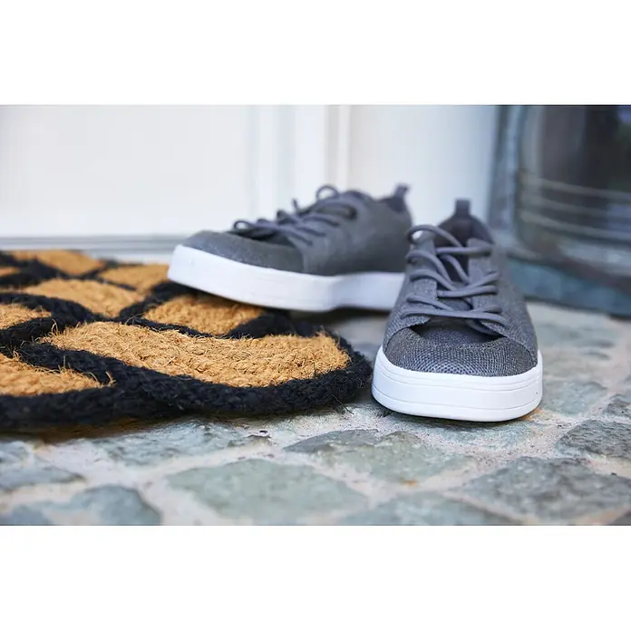 Fußmatte Nature Classic - 75 x 45 cm, Natur/Schwarz, Material Nutzschicht: 100 % Kokos - Ein Paar graue Textil-Sneaker mit weißen Sohlen liegt auf einer braun-schwarz gemusterten Fußmatte vor einer Tür.