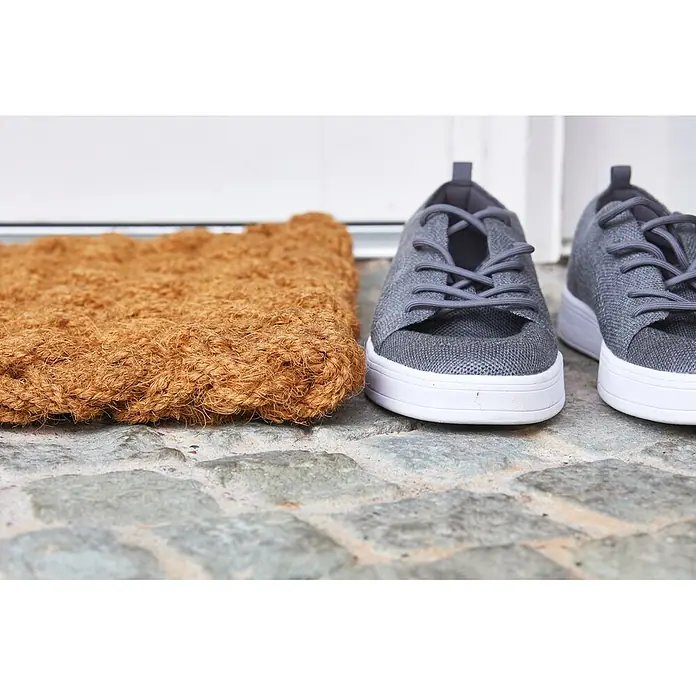 Fußmatte Nature Classic - 75 x 45 cm, Natur, Material Nutzschicht: 100 % Kokos, Karo - Eine braune Kokosfußmatte und ein Paar graue Strick-Sneaker mit weißen Sohlen liegen auf einer Steinterrasse vor einer Haustür.