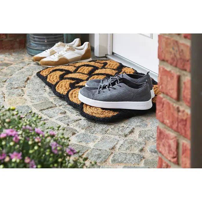 Fußmatte Nature Classic - 75 x 45 cm, Natur/Schwarz, Material Nutzschicht: 100 % Kokos - Graue Strickschuhe mit weißen Sohlen auf einer gemusterten Fußmatte vor einer Tür. Beige Sneaker sind im Hintergrund.