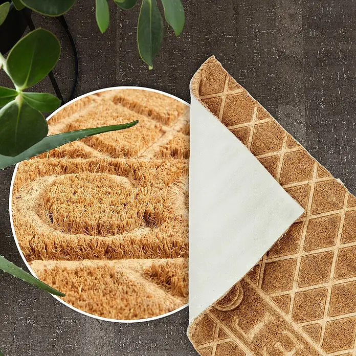 Fußmatte Coco Shape - 75 x 45 cm, Natur, Material Nutzschicht: 100 % Kokos, Welcome - Braune Kokos-Fußmatte mit Herzmuster, teilweise umgeklappt, zeigt strukturierte Unterseite und weiße Rückseite, auf dunklem Boden mit Pflanzen.