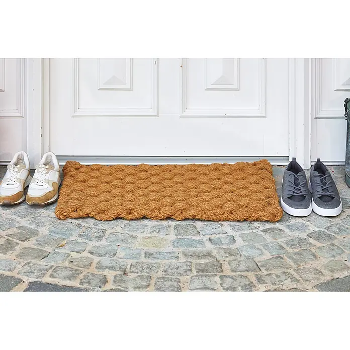 Fußmatte Nature Classic - 75 x 45 cm, Natur, Material Nutzschicht: 100 % Kokos, Karo - Braune Kokosfasermatte vor einer weißen Tür auf Kopfsteinpflaster, flankiert von zwei Paar Schuhen.