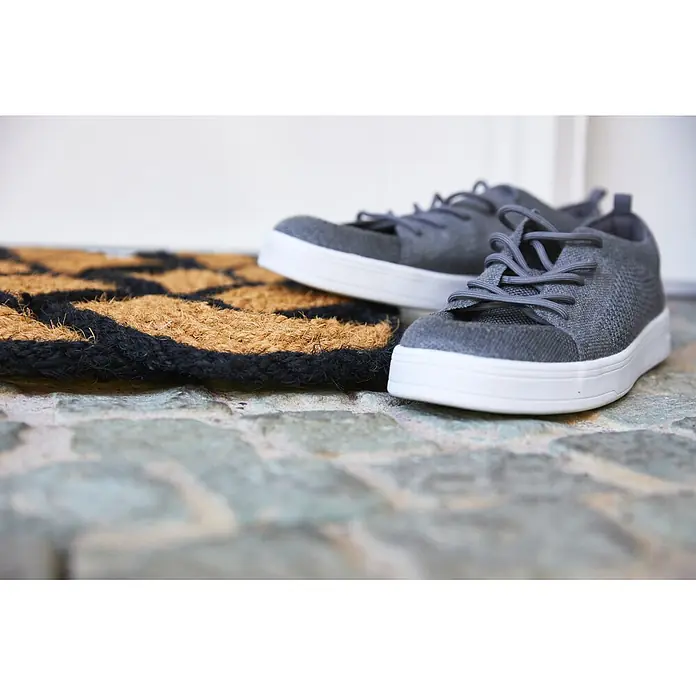 Fußmatte Nature Classic - 75 x 45 cm, Natur/Schwarz, Material Nutzschicht: 100 % Kokos - Ein Paar graue Sneaker mit weißen Sohlen ruht auf einer schwarz-braun gemusterten Fußmatte auf einem Steinboden.