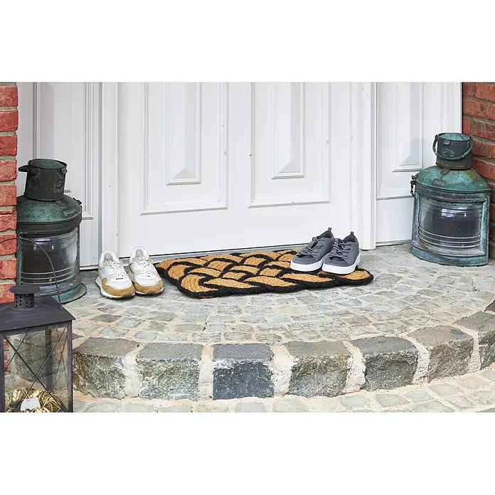 Fußmatte Nature Classic - 75 x 45 cm, Natur/Schwarz, Material Nutzschicht: 100 % Kokos - Geflochtene Fußmatte aus Naturfaser mit schwarz-beigem Knotenmuster vor einer Haustür mit Schuhen.
