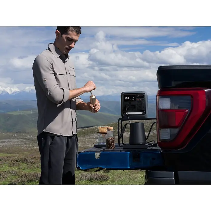DJI Powerstation Power 1000 Mini - 1.008 Wh, Schwarz - Graue tragbare Powerstation auf schwarzem Ständer, versorgt einen Wasserkocher für die Kaffeezubereitung im Freien, neben einem Mann, der Kaffee mahlt.