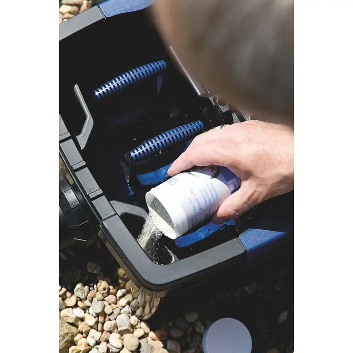 Oase AquaActiv Filterstarter AquaActiv - 200 ml - Eine Hand schüttet granulierten Teichfilterstarter aus einem weißen Behälter in einen schwarzen Filterkasten mit blauen Elementen.