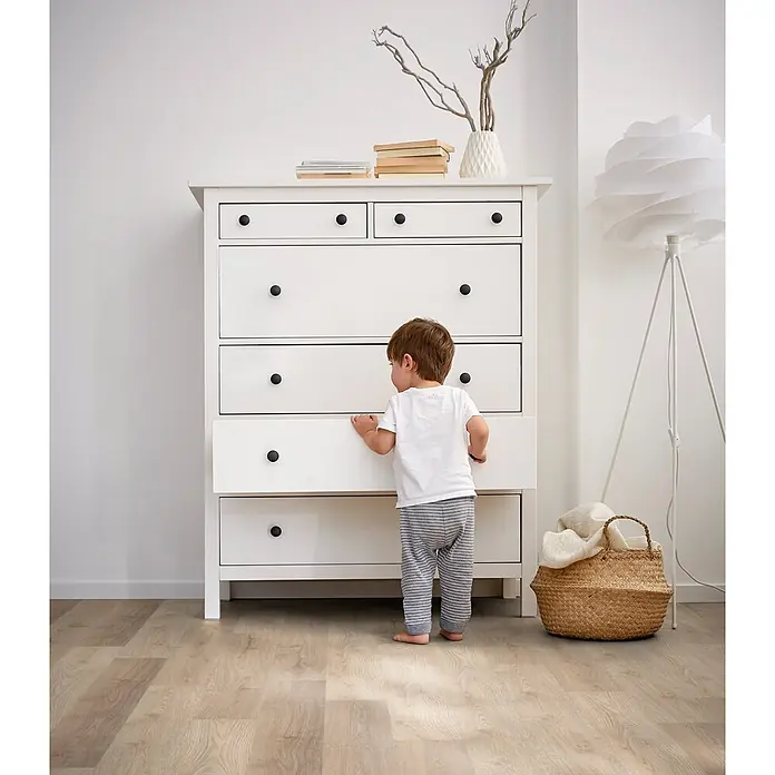 Decolife Designboden Freestyle Access Hardy Oak - 1.220 x 185 x 8,5 mm, Landhausdiele, Hardy Oak - Weiße Kommode mit schwarzen Knöpfen. Ein Kleinkind zieht eine der unteren Schubladen auf, was die Nutzung im Kinderzimmer zeigt.