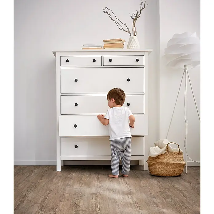 Decolife Designboden Freestyle Access Tuscan Pine - 1.220 x 185 x 8,5 mm, Landhausdiele, Tuscan Pine - Weiße Kommode mit schwarzen Knaufgriffen, ein Kind öffnet eine untere Schublade.