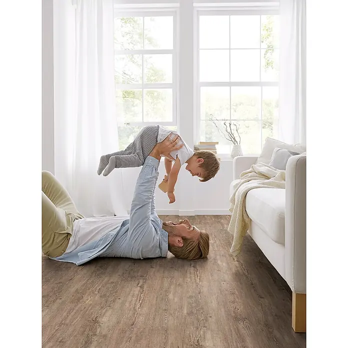 Decolife Designboden Freestyle Access Tuscan Pine - 1.220 x 185 x 8,5 mm, Landhausdiele, Tuscan Pine - Warmbrauner Laminatboden in Holzoptik in einem hellen Wohnzimmer mit spielendem Vater und Kind.