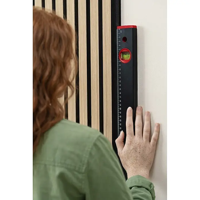 Alpha Tools Set de niveles de burbuja - 3 pzs. - Nivel de burbuja rojo y negro con regla, utilizado por una persona para comprobar la alineación vertical en una pared con listones de madera.