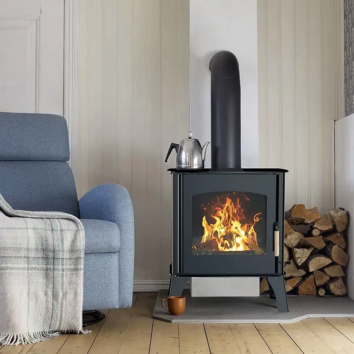 Sannover Kaminofen - 11,8 kW, Raumheizvermögen: 240 m³, Material Verkleidung: Stahl, Schwarz - Schwarzer Holzofen mit sichtbarem Feuer, einem Wasserkessel darauf und gestapeltem Brennholz, der eine gemütliche Atmosphäre im Wohnzimmer schafft.