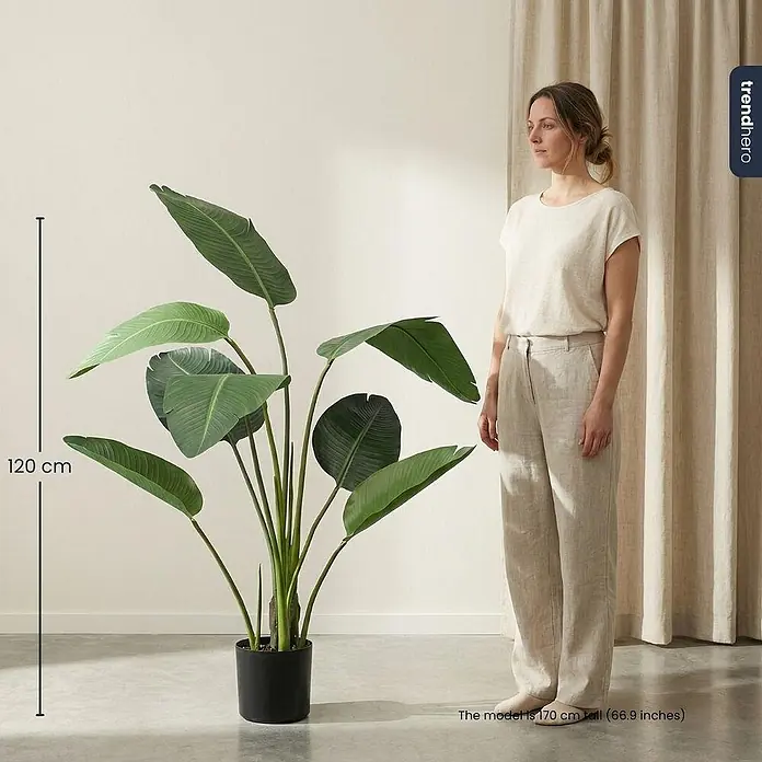 Florora Kunstpflanze Strelitzia - Höhe: 120 cm, Holz - Große künstliche Strelitzienpflanze mit grünen Blättern in schwarzem Topf, 120 cm hoch, zum Größenvergleich neben einer Person.