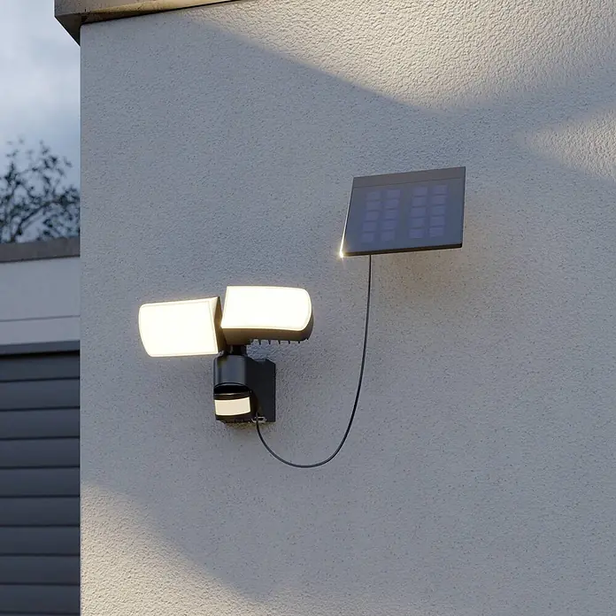 Arcchio Außenwandleuchte Lissano - schwarz, IP44 - Dunkelgrauer Solar-LED-Fluter mit zwei verstellbaren Köpfen und Bewegungssensor, verbunden mit separatem Solarpanel, beleuchtet eine strukturierte Wand.