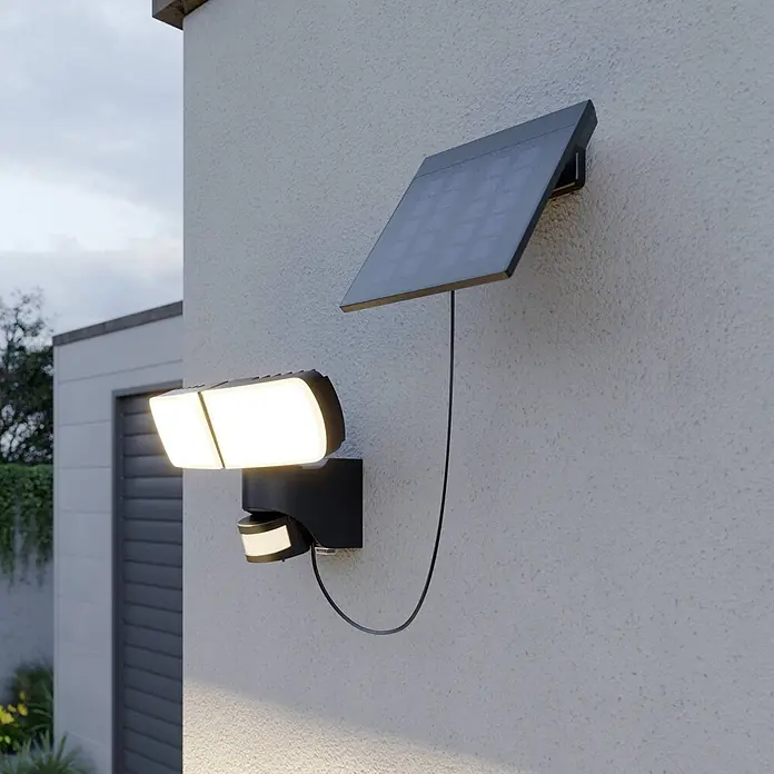Arcchio Außenwandleuchte Lissano - schwarz, IP44 - Schwarze Solar-Sicherheitsleuchte mit zwei LED-Panels und Bewegungssensor, verbunden mit einem Solarpanel, an einer Außenwand montiert, leuchtend.