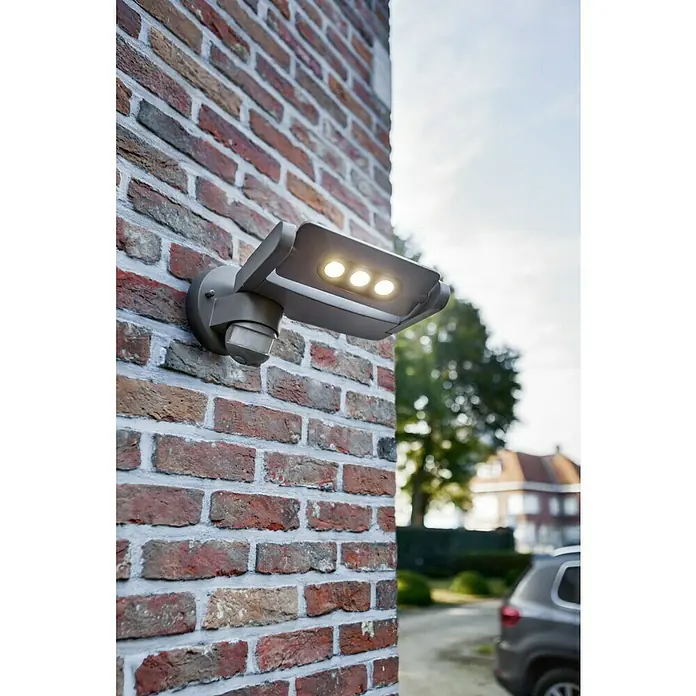 Lutec Aplique LED con sensor - 9 W, L x An x Al: 23 x 23 x 13,5 cm, Gris, Blanco neutro - Una luz exterior gris con sensor de movimiento está montada en una pared de ladrillos, proporcionando una iluminación brillante.