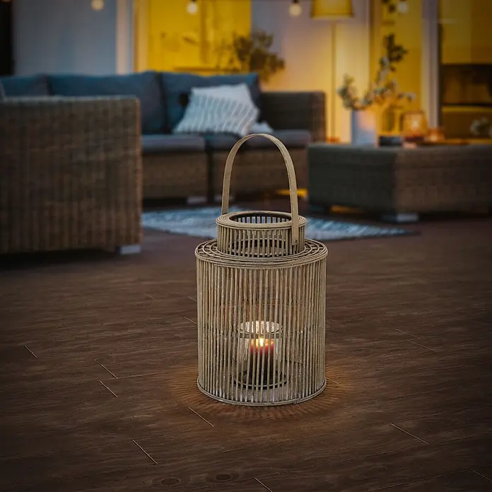 ECD Germany Windlicht Boho Bambus Laterne - Ø x H: 28,5 x 54,5 cm, Natur, 1 Stk. - Laterne aus Naturholz mit brennender Kerze, die einen warmen Schein auf einen dunklen Terrassenboden wirft, mit unscharfen Gartenmöbeln im Hintergrund.