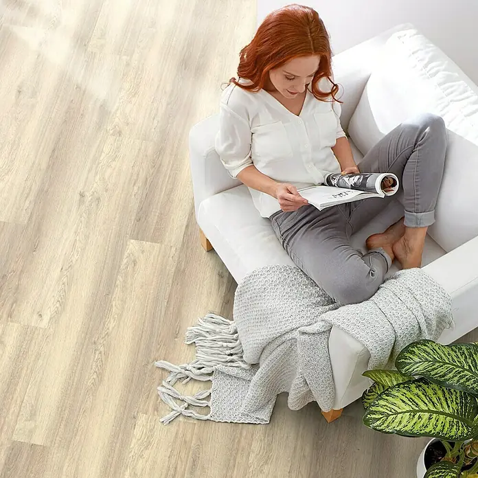 Decolife Designboden - Oak Baltic, Landhausdiele, 1.220 x 185 x 10,5 mm - Helle Holzbodenfliesen schaffen eine warme und einladende Atmosphäre, während eine Frau entspannt auf einem weißen Sessel ein Magazin liest.