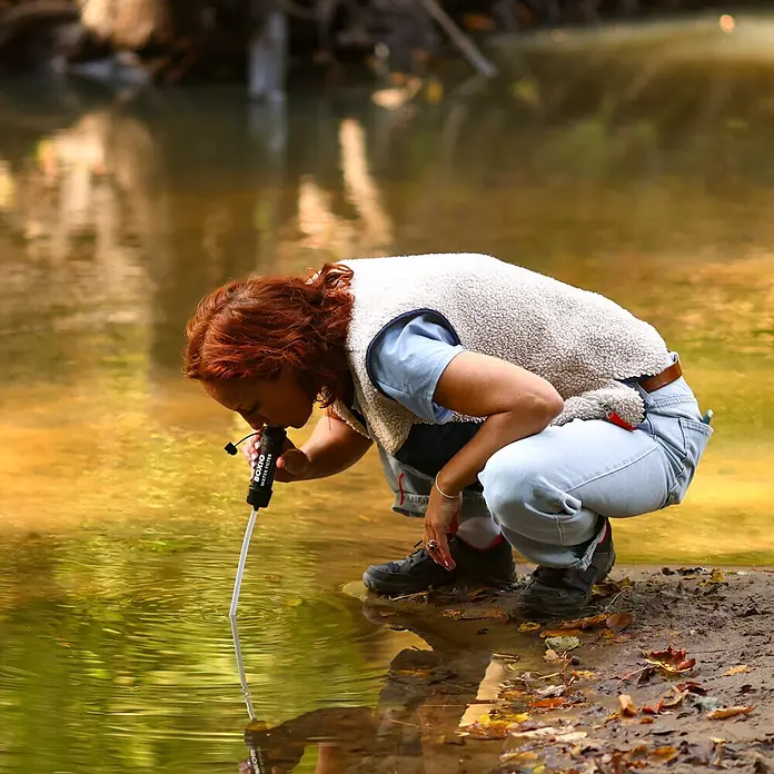 Boxio Wasserfilter - 3 -tlg. - Schwarzer BOXIO WASSERFILTER mit weißem Strohhalm, genutzt von Person zum Trinken direkt aus einem Bach.