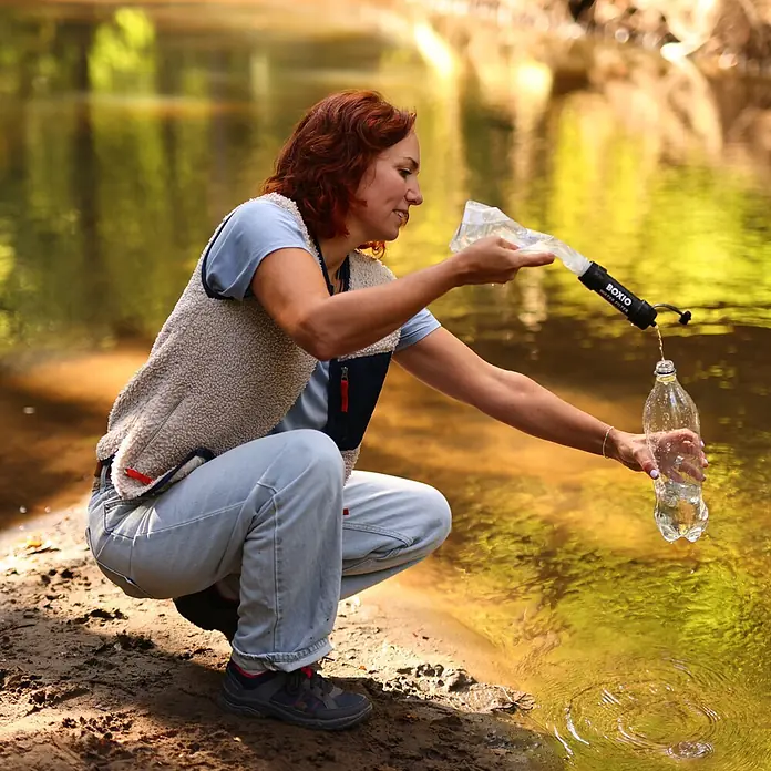 Boxio Wasserfilter Set - 5 -tlg. - Eine Frau filtert Flusswasser mit einem schwarzen BOXIO Wasserfilter in eine Flasche.