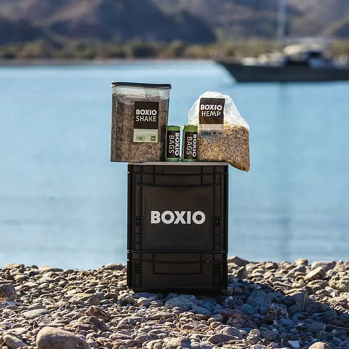 Boxio Trenntoiletten-Streu - Passend für: Boxio Campingtoilette - BOXIO Produkte (Shake, Hanfstreu, Beutel) auf einer schwarzen BOXIO Kiste am Felsstrand am Meer.