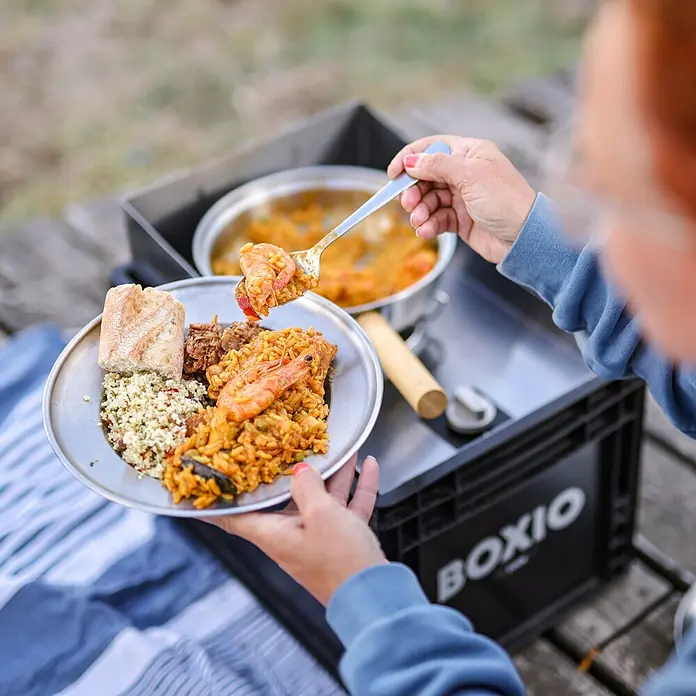Boxio Gaskocher Cook Plus - 40 x 30 x 27 cm, Edelstahl - Person serviert Paella von einem Topf auf einem schwarzen BOXIO Outdoor-Kochsystem. Essen beinhaltet Paella, Couscous und Brot auf einem Metallteller.