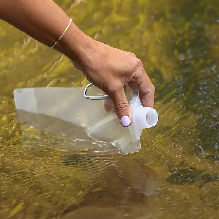 Boxio Wasserfilter - 3 -tlg. - Eine Hand füllt einen transparenten, faltbaren Wasserbeutel aus einem Bach. Der Beutel hat einen weißen Verschluss und einen Karabinerhaken.