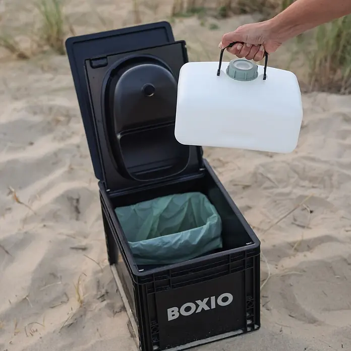 Boxio Müllbeutel BioBags - 10 l, Grün - Ein offenes schwarzes Trockenklo mit grünem Beutel steht am Sandstrand. Eine Hand hält einen weißen Wasser-/Abfallbehälter darüber.