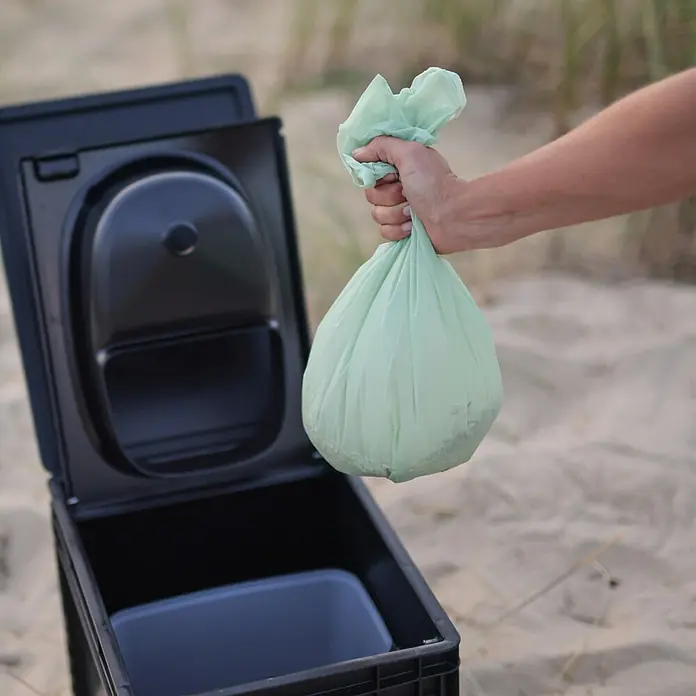 Boxio Müllbeutel BioBags - 10 l, Grün - Hand hält grünen Müllbeutel über offener schwarzer Campingtoilette.