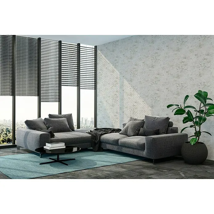 A.S. Création New Walls Vliestapete Used Wall - Grau, Betonoptik, 10,05 x 0,53 m - Großes graues Ecksofa mit stilvollem Couchtisch auf einem blauen Teppich, vor Fenstern mit Jalousien und einer Zimmerpflanze.