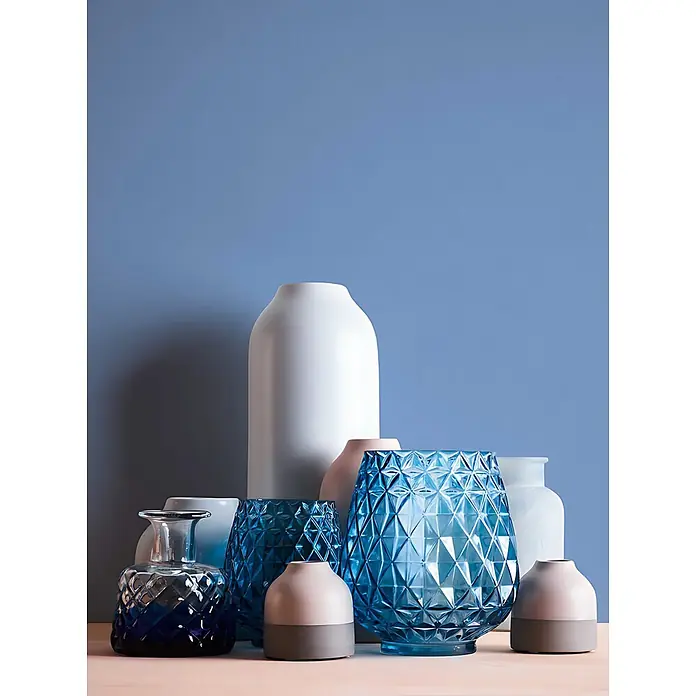 Profhome Vliestapete Vliestapete Uni in blau - Uni, 10.050 x 530 mm - Eine Ansammlung von Glas- und Keramikvasen und Kerzenhaltern in Blau, Weiß, Pink und Grau, arrangiert auf heller Oberfläche vor blauer Wand.