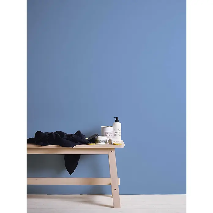 Profhome Vliestapete Vliestapete Uni in blau - Uni, 10.050 x 530 mm - Helle Holzbank mit schwarzem Textil und Beauty-Produkten darauf, vor einer blauen Wand.