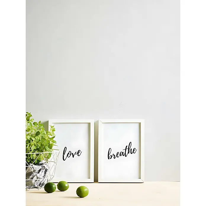 Profhome Vliestapete Vliestapete Uni in weiß - Uni, 10.050 x 530 mm - Zwei weiße gerahmte Bilder mit 'love' und 'breathe', mit einer Pflanze und Limetten auf einer Holzoberfläche.