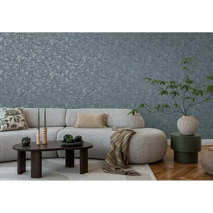 Profhome Vliestapete Heißgeprägte Vliestapete Ton-In-Ton, Floral, Textiloptik in blau - blau-grauplatin, 10.050 x 530 mm - Grünblaue Tapete mit dezentem Blattmuster, die eine Wohnzimmerwand hinter einem hellen Sofa schmückt.