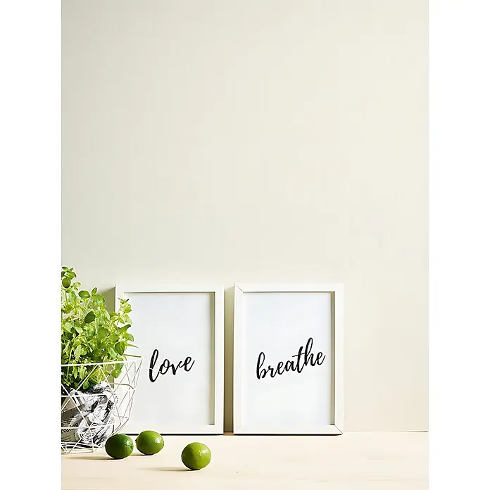 Profhome Vliestapete Vliestapete zum Überstreichen Textiloptik in weiß - 21.000 x 1.060 mm - Zwei weiße gerahmte Drucke mit „love“ und „breathe“ in schwarzer Schrift, daneben eine Topfpflanze und Limetten auf Holzoberfläche.