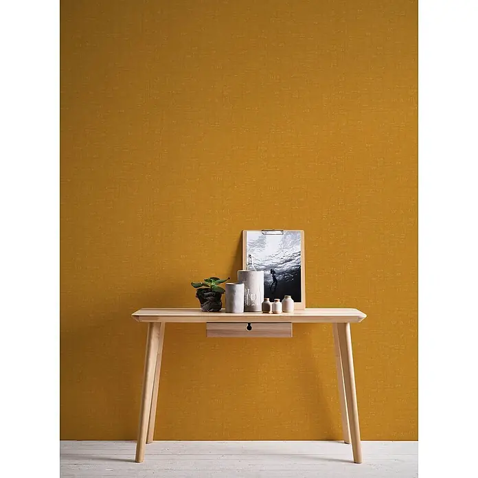 Profhome Vliestapete Heißgeprägte Vliestapete Textiloptik in gelb - ocker-gelb, 10.050 x 530 mm - Heller Konsolentisch aus Holz mit Dekorationsartikeln, Pflanze und Bild, vor einer strukturierten senfgelben Wand.